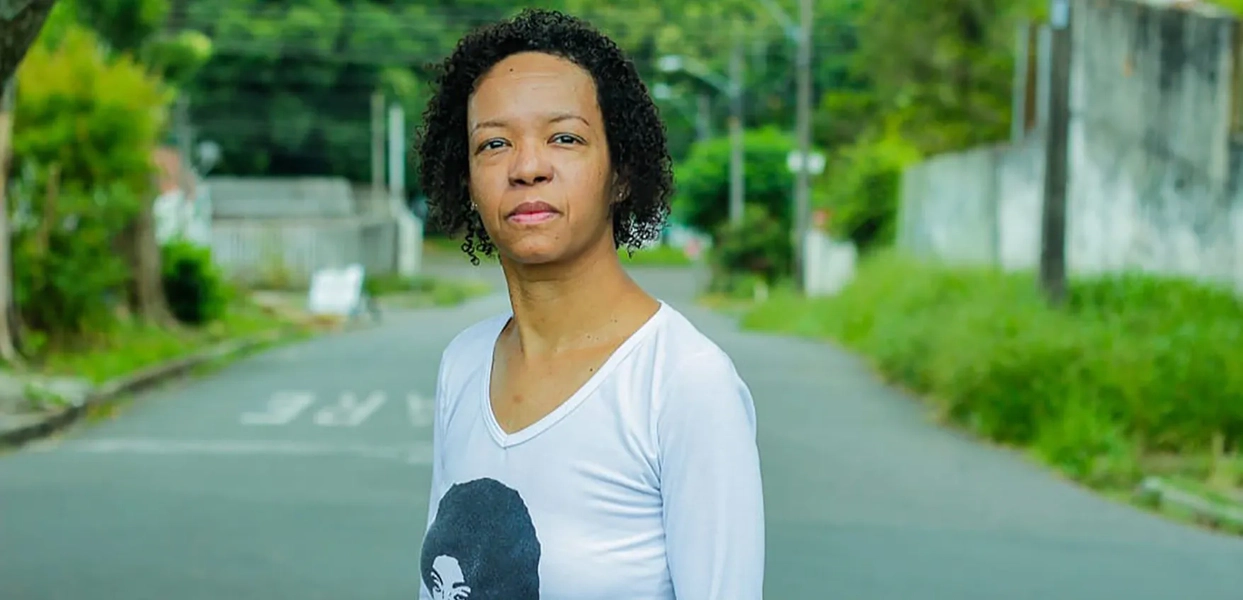 Silvanna Bárbara, professora do Escola Nota 10, com uma blusa de manga longa branca e cabelo cacheado curto