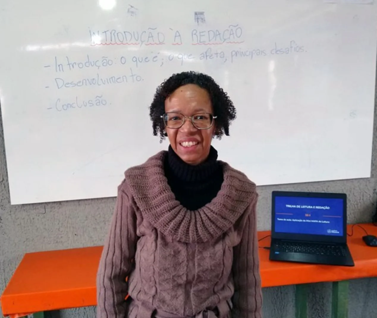 Silvana Bárbara de blusa de frio cinza em frente a uma mesa laranja e um quadro escolar branco