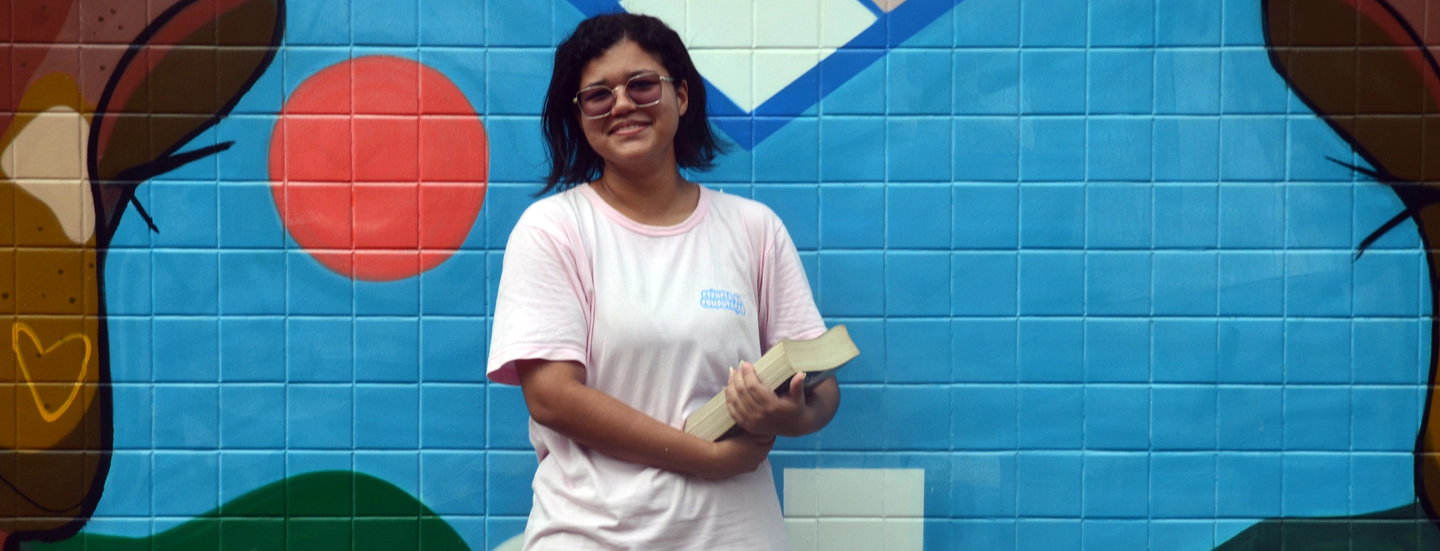 Marina Santos, de blusa branca e calça preta  sorrindo segurando um livro 